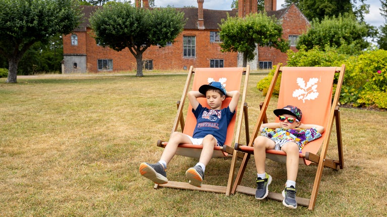 Two children relaxing in National Trust deckchairs on the lawn at Baddesley Clinton, Warwickshire